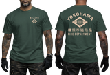 Front and back views of a dark green short-sleeve t-shirt worn by a model. The front features a small beige chest logo, and the back displays 'YOKOHAMA' in arched beige letters above a diamond emblem and 'FIRE DEPARTMENT' text.