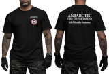 Two-panel photo showing the front and back of a black short-sleeve t-shirt. The front features a small circular "Antarctic Fire Department McMurdo Station" logo on the left chest. The back displays large white text: "ANTARCTIC FIRE DEPARTMENT McMurdo Station
