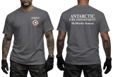 Two-panel photo showing the front and back of a charcoal short-sleeve t-shirt. The front features a small circular "Antarctic Fire Department McMurdo Station" logo on the left chest. The back displays large white text: "ANTARCTIC FIRE DEPARTMENT McMurdo Station
