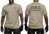 Two-panel photo showing the front and back of a sand short-sleeve t-shirt. The front features a small circular "Antarctic Fire Department McMurdo Station" logo on the left chest. The back displays large white text: "ANTARCTIC FIRE DEPARTMENT McMurdo Station