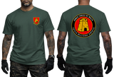 A man modeling a dark green t-shirt, showing both the front and back views. The front features a small red and yellow military crest on the left chest.