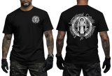 Two-view composite image of a black t-shirt displayed on a male model with tattooed arms and tactical gloves. The left side shows the front of the shirt with a small left-chest logo. The right side shows the back of the shirt with a large, detailed white graphic print of the Saint Benedict Medal and the circular text 'INTERNATIONAL CATHOLIC ASSOCIATION of EXORCISTS'.