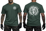 Two-view composite image of a forest green t-shirt displayed on a male model with tattooed arms and tactical gloves. The left side shows the front of the shirt with a small left-chest logo. The right side shows the back of the shirt with a large, detailed white graphic print of the Saint Benedict Medal and the circular text 'INTERNATIONAL CATHOLIC ASSOCIATION of EXORCISTS'.