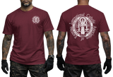 Two-view composite image of a maroon t-shirt displayed on a male model with tattooed arms and tactical gloves. The left side shows the front of the shirt with a small left-chest logo. The right side shows the back of the shirt with a large, detailed white graphic print of the Saint Benedict Medal and the circular text 'INTERNATIONAL CATHOLIC ASSOCIATION of EXORCISTS'.