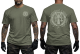 Two-view composite image of a military green t-shirt displayed on a male model with tattooed arms and tactical gloves. The left side shows the front of the shirt with a small left-chest logo. The right side shows the back of the shirt with a large, detailed white graphic print of the Saint Benedict Medal and the circular text 'INTERNATIONAL CATHOLIC ASSOCIATION of EXORCISTS'.