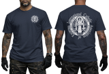 Two-view composite image of a navy t-shirt displayed on a male model with tattooed arms and tactical gloves. The left side shows the front of the shirt with a small left-chest logo. The right side shows the back of the shirt with a large, detailed white graphic print of the Saint Benedict Medal and the circular text 'INTERNATIONAL CATHOLIC ASSOCIATION of EXORCISTS'.