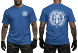 Two-view composite image of a royal blue t-shirt displayed on a male model with tattooed arms and tactical gloves. The left side shows the front of the shirt with a small left-chest logo. The right side shows the back of the shirt with a large, detailed white graphic print of the Saint Benedict Medal and the circular text 'INTERNATIONAL CATHOLIC ASSOCIATION of EXORCISTS'.